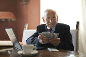 Confident senior businessman holding money in hands while sitting at table near laptop